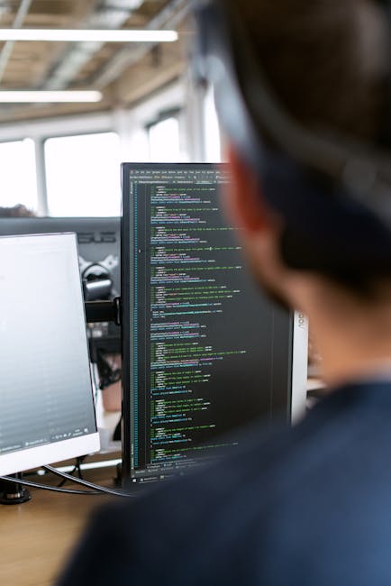A person working in a modern office environment, viewed from behind, focuses on multiple computer screens displaying lines of code with colorful syntax highlighting. The workspace features a large win